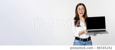 Young woman in glasses showing laptop screen, demonstrating promo on computer, website or store, standing over white background 96585242