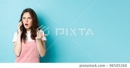 Portrait of confused and shocked woman receive bad news while talking on smartphone, receive bad phone call, standing against blue background 96585268