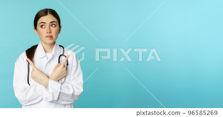 Portrait of indecisive, confused female doctor, pharmacy worker pointing sideways and biting lip clueless, dont know, standing over blue background 96585269