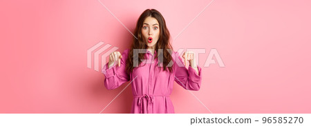Stylish caucasian woman saying wow, pointing fingers down at impressive banner news, showing advertisement, staring amazed, standing against pink background 96585270
