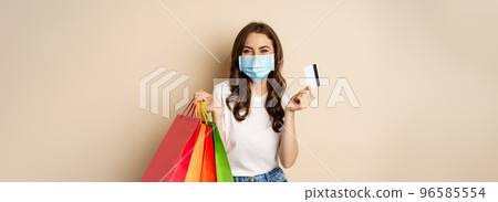 Covid-19 pandemic and lifestyle concept. Young woman posing in medical face mask with shopping bags from mall, holding credt card in hand, beige background Covid-19 pandemic and lifestyle concept. Young woman posing in medical face mask with shopping bags from mall, holding credt card in hand, beige background 96585554