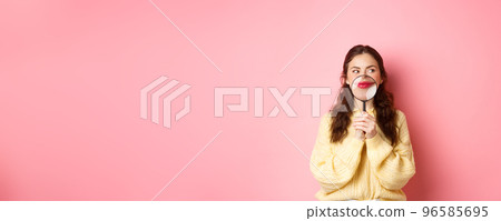 Image of coy attractive girl thinking of something interesting, showing her smile with magnifying glass near lips, standing against pink background, looking aside at promotional text 96585695