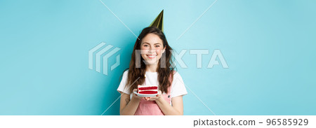 Holidays and celebration. Cheerful birthday girl in party hat holding bday cake and smiling, making wish on lit candle, standing against blue background 96585929