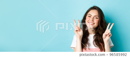 Close up portrait of attractive positive girl saying cheeze, smiling and showing v-signs peace, standing over blue background Close up portrait of attractive positive girl saying cheeze, smiling and showing v-signs peace, standing over blue background 96585992