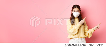 Covid, corona and social distancing concept. Excited female model in respirator shows you advertisement, points at logo aside, wears medical respirator on face, pink background Covid, corona and social distancing concept. Excited female model in respirator shows you advertisement, points at logo aside, wears medical respirator on face, pink background 96585994