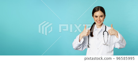 Happy woman doctor, medical worker in white coat, showing thumbs up in approval, like something, praise, standing over blue background 96585995