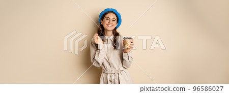 Beautiful happy girl drinking takeaway coffee from cafe and smiling, posing with cup of beverage, beige background 96586027