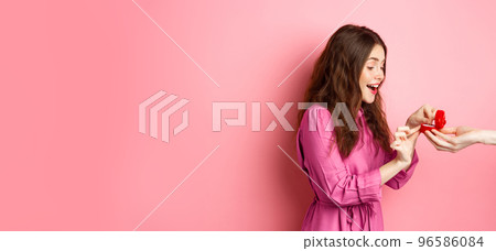 Portrait of surprised girlfriend receive marriage proposal, looking at hand with engagement ring and scream of surprise and excitement, standing over pink background Portrait of surprised girlfriend receive marriage proposal, looking at hand with engagement ring and scream of surprise and excitement, standing over pink background 96586084