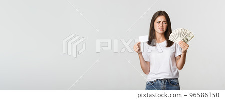 Confused and displeased young woman looking at money and frowning, prefer credit card 96586150