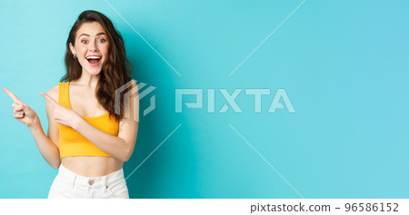 Portrait of happy young woman smiling amused, showing awesome promo deal, pointing fingers left at copy space and looking fascinated at camera, standing in summer clothes against blue background 96586152