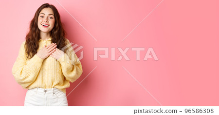 Portrait of cute heartfelt girl appreciate nice gesture, being praised, holding hands on heart and sighing from romantic feelings, looking grateful, say thank you, standing over pink background 96586308