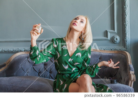 Beautiful blonde woman in green dress sitting on blue sofa using perfume Beautiful blonde woman in green dress sitting on blue sofa using perfume 96586339