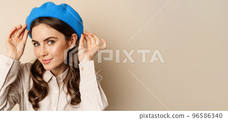 Stylish modern girl put on trendy hat on head and smiling, going out, posing against beige background Stylish modern girl put on trendy hat on head and smiling, going out, posing against beige background 96586340