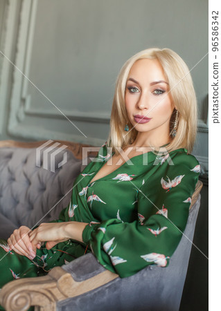 Beautiful blond woman with makeup in green dress sitting on grey velvet sofa Beautiful blond woman with makeup in green dress sitting on grey velvet sofa 96586342