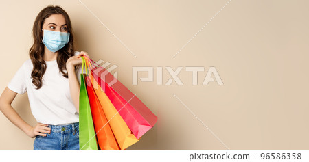 Covid-19 pandemic and lifestyle concept. Young woman posing in medical face mask with shopping bags from mall, vaccinated girl going in store in personal protective equipment, beige background Covid-19 pandemic and lifestyle concept. Young woman posing in medical face mask with shopping bags from mall, vaccinated girl going in store in personal protective equipment, beige background 96586358