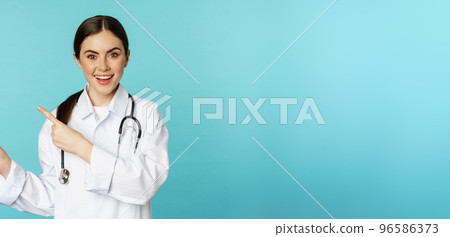 Portrait of smiling medical worker, girl doctor in white coat with stethoscope, pointing fingers left, showing medical clinic advertisement, torquoise background Portrait of smiling medical worker, girl doctor in white coat with stethoscope, pointing fingers left, showing medical clinic advertisement, torquoise background 96586373