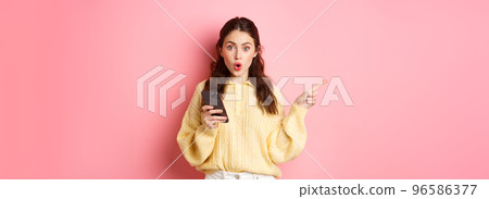 Surprised young beautiful girl gasping and saying wow, pointing finger aside at left copyspace, holding mobile phone, standing against pink background Surprised young beautiful girl gasping and saying wow, pointing finger aside at left copyspace, holding mobile phone, standing against pink background 96586377