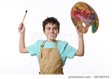 Inspired smiling teenage boy - talented painter in blue t-shirt and beige apron holding paintbrush and wooden palette, Development of children's creativity and drawing with paints. Art. Entertainment 96586503