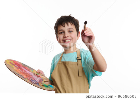 Artistic teenage boy wearing beige apron, holding a paintbrush and a color palette, paints on imaginary canvas, looking at camera, isolated on white background. Kids entertainment and art development 96586505