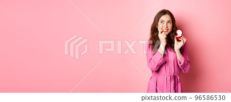 Beautiful girl dream of her wedding day, looking thoughtful up, showing engagement ring in red box and smiling, standing over pink background Beautiful girl dream of her wedding day, looking thoughtful up, showing engagement ring in red box and smiling, standing over pink background 96586530