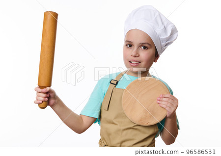 Adorable Caucasian child boy, wearing a chef's hat and beige apron, posing with a rolling pin and wooden cutting board, isolated on white background. Baker Confectioner. Chef pastry. Cook profession Adorable Caucasian child boy, wearing a chef's hat and beige apron, posing with a rolling pin and wooden cutting board, isolated on white background. Baker Confectioner. Chef pastry. Cook profession 96586531