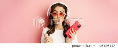 Stylish young woman with lolipop and cellphone, wearing sunglasses and headphones, listening music, standing over pink background 96586552