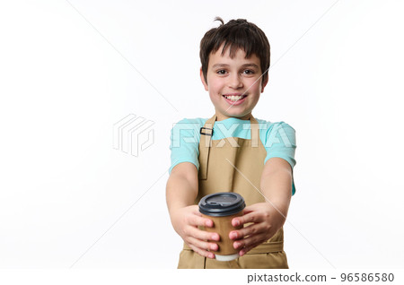 Adorable happy teenage boy wearing blue t-shirt and beige chef's apron, smiling a toothy smile to camera, handing out an eco-friendly cardboard mug of takeaway coffee, isolated over white background 96586580