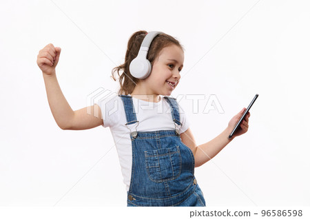 Adorable happy Caucasian little girl wearing blue denim overalls, holding mobile phone in her hands, dances and listens to the music on her stereo headphones, isolated white background, copy ad space Adorable happy Caucasian little girl wearing blue denim overalls, holding mobile phone in her hands, dances and listens to the music on her stereo headphones, isolated white background, copy ad space 96586598