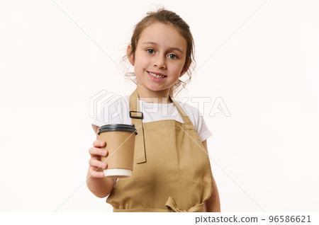 Beautiful blue-eyed Caucasian little girl wearing a beige chef's apron, handing out a takeaway hot beverage, tea or coffee drink in an ecologically-friendly paper cup, cutely smiles looking at camera 96586621