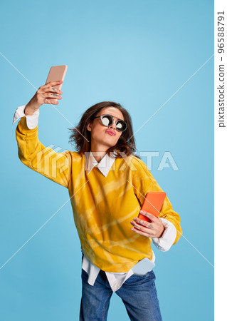 Portrait of young beautiful girl posing in sunglasses and yellow sweater, taking selfie with two phones isolated over blue background Portrait of young beautiful girl posing in sunglasses and yellow sweater, taking selfie with two phones isolated over blue background 96588791