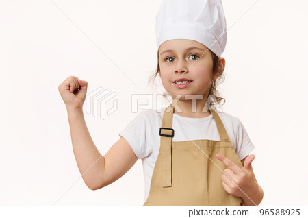 European beautiful little girl - chef pastry, cook, baker confectioner wearing kitchen apron and hat, isolated on white background with free space for promotional text. Cooking class for talented kids 96588925