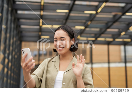 Happy asian girl waves at her smartphone camera, says hello to friend on video chat, calling someone, standing on street Happy asian girl waves at her smartphone camera, says hello to friend on video chat, calling someone, standing on street 96589739