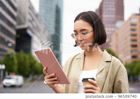 Portrait of happy young woman in glasses, standing on street with cup of coffee and tablet 96589740