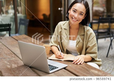Portrait of young woman sitting in cafe with laptop, making notes, smiling at camera, working, having online meeting Portrait of young woman sitting in cafe with laptop, making notes, smiling at camera, working, having online meeting 96589741