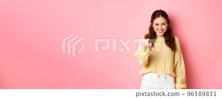 Young confident and determined woman smiling, pointing finger at camera, making her choise, inviting you, standing against pink background Young confident and determined woman smiling, pointing finger at camera, making her choise, inviting you, standing against pink background 96589831