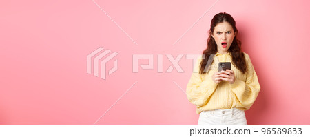 Confused and angry girlfriend reading strange message on phone, staring at camera frustrated and questioned, standing against pink background Confused and angry girlfriend reading strange message on phone, staring at camera frustrated and questioned, standing against pink background 96589833