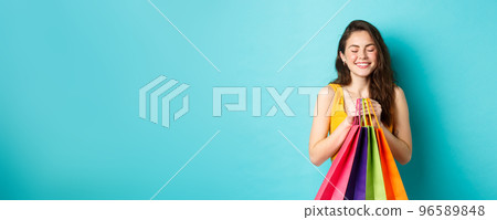 Young beautiful woman daydreaming about wearing new clothes, holding shopping bags, close eyes and smile dreamy, standing over blue background 96589848
