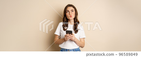 Portrait of woman thinking, holding mobile phone and looking up with thoughtful face, standing over beige background 96589849