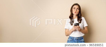 Portrait of woman thinking, holding mobile phone and looking up with thoughtful face, standing over beige background Portrait of woman thinking, holding mobile phone and looking up with thoughtful face, standing over beige background 96589851
