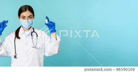 Smiling medical staff, doctor in face mask and rubber gloves, showing syringe and vaccine from covid-19 pandemic, standing over blue background Smiling medical staff, doctor in face mask and rubber gloves, showing syringe and vaccine from covid-19 pandemic, standing over blue background 96590109