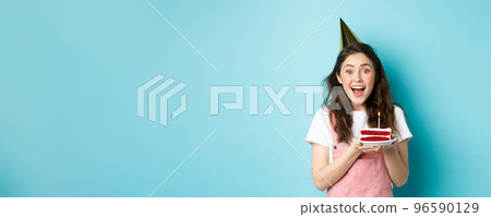 Holidays and celebration. Cheerful birthday girl in party hat holding bday cake and smiling, making wish on lit candle, standing against blue background 96590129