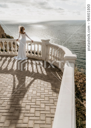 Romantic bride, blond girl in white wedding dress with open shoulders posing on open terrace with backdrop of the sea and rocks. Stylish young woman standing on terrace and looking on ocean at sunset Romantic bride, blond girl in white wedding dress with open shoulders posing on open terrace with backdrop of the sea and rocks. Stylish young woman standing on terrace and looking on ocean at sunset 96601800