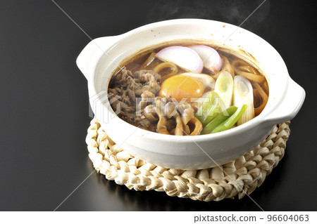 Hot miso stewed udon with beef, shimeji mushrooms, green onion, kamaboko, and egg yolk photographed against a black background 96604063