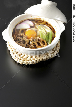 Hot miso stewed udon with beef, shimeji mushrooms, green onion, kamaboko, and egg yolk photographed against a black background 96604081