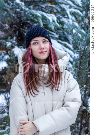 Beautiful attractive young woman in fashionable clothes in winter. Portrait of a girl with long pink hair in a hat and jacket, it is snowing. Beautiful attractive young woman in fashionable clothes in winter. Portrait of a girl with long pink hair in a hat and jacket, it is snowing. 96605314