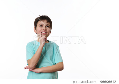Happy teenage boy wearing turquoise t-shirt, holding his hand at chin and cutely smiling, thoughtfully looking at aside, isolated over white background with copy ad space for promotional text 96606612