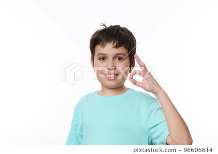 Dark-haired Caucasian boy in blue turquoise casual t-shirt, smiling a cheerful beautiful toothy smile, showing ok sign looking at camera, isolated over white background with space for promotional text 96606614