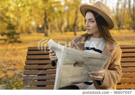 An attractive girl reads a newspaper in the park. Surveillance private detective monitors the object.Attractive intelligence agent An attractive girl reads a newspaper in the park. Surveillance private detective monitors the object.Attractive intelligence agent 96607097