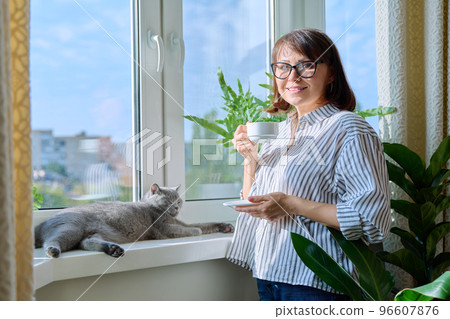 Middle aged woman with cup of coffee and pet, looking at camera near window Middle aged woman with cup of coffee and pet, looking at camera near window 96607876