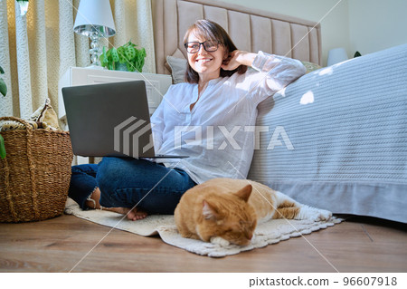 Middle aged smiling woman sitting on floor at home using laptop with sleeping cat Middle aged smiling woman sitting on floor at home using laptop with sleeping cat 96607918